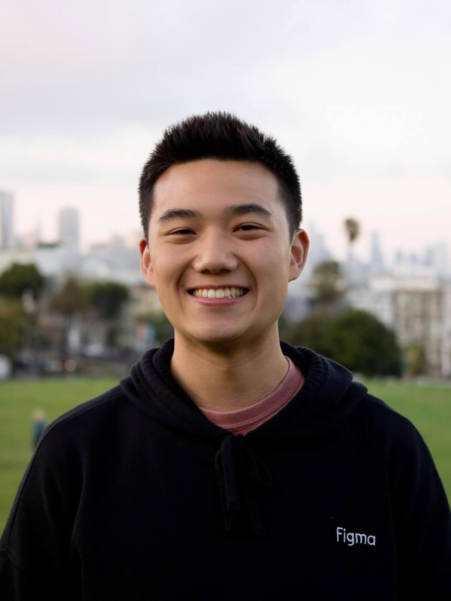 Austin Lee, smiling in a black hoodie, stands in a park with a blurred city skyline behind him.